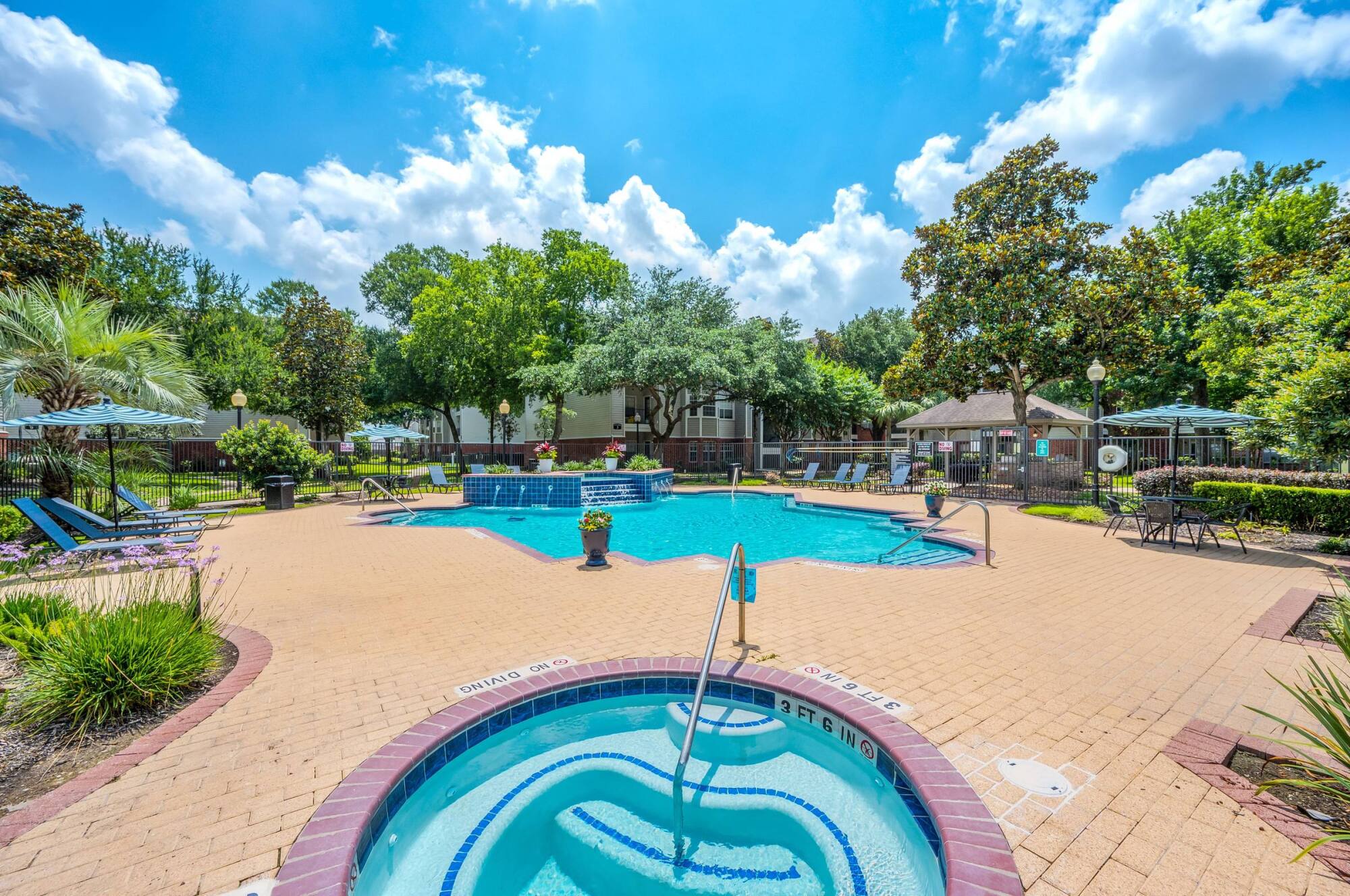 The spa and sparkling swimming pool at Vinings in Stafford, TX, surrounded by a paver sundeck, lounge chairs, and lush trees.