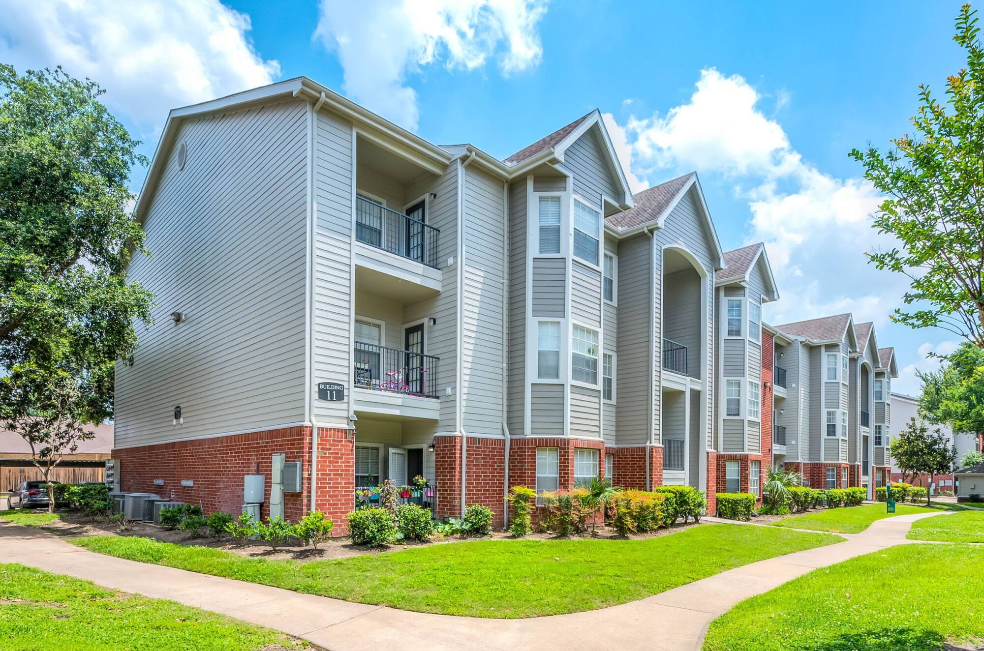 The three-story brick and siding apartment buildings at Vinings in Stafford, TX, with private balconies, green lawns, and a winding sidewalk.